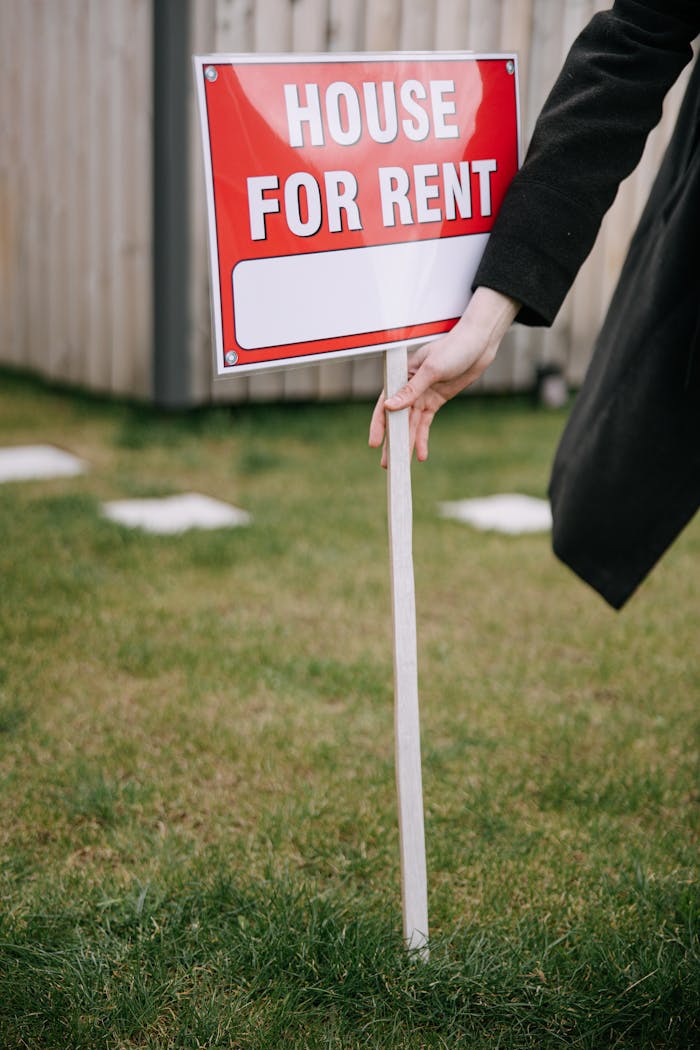 Close-up of a red House for Rent sign on a grassy lawn, hand holding pole.