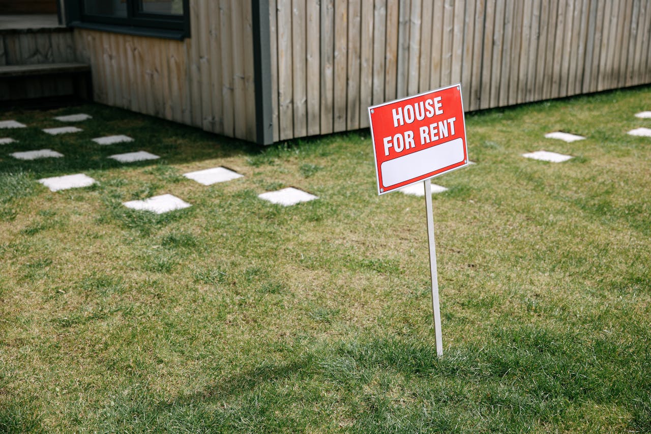 about-01 A red House for Rent sign stands on a grassy lawn beside a wooden house exterior.